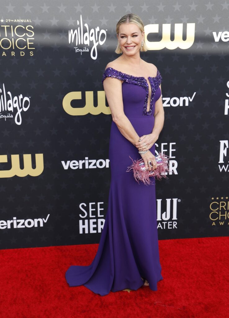 A woman in a purple gown with embellishments poses confidently on the red carpet at an awards event.
